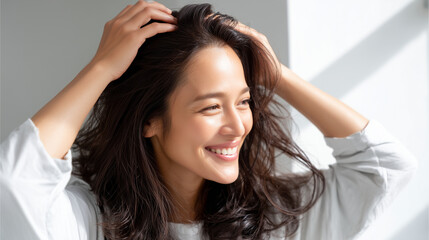 Naklejka premium A positive, high-key commercial shot of a smiling Japanese woman with healthy, voluminous dark hair, gently massaging her scalp with her fingertips in a sunlit room. Hair recovery,