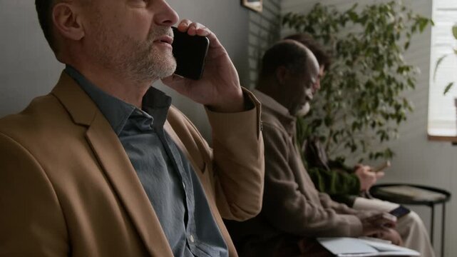 Close up tilt up shot of middle aged businessman talking on phone and holding documents while sitting in bank waiting area