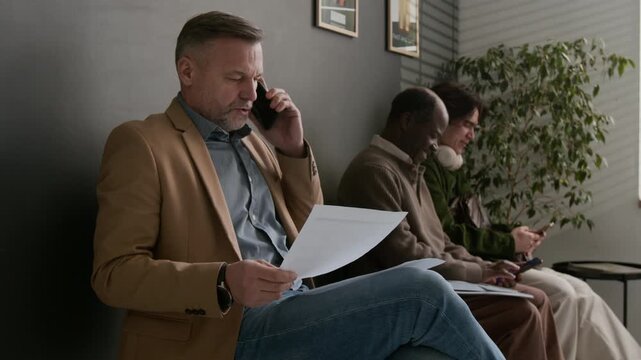 Middle aged man in blazer, talking on phone with papers in his hand, sitting in waiting area of modern bank