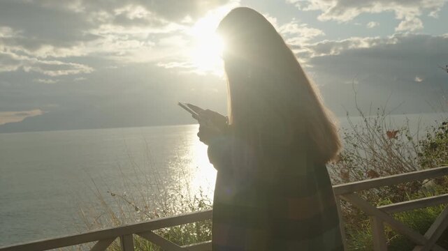 A Woman at the Edge of a Cliff by the Sea, with Mountains and the Sun's Rays on the Water in the Background, Looks at Her Phone.
