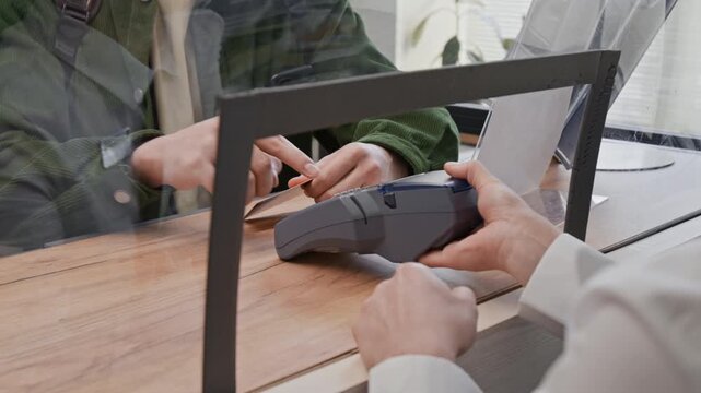 Close up over the shoulder shot of unrecognizable customer usinng credit card on terminal, while bank teller holding device across protective glass barrier