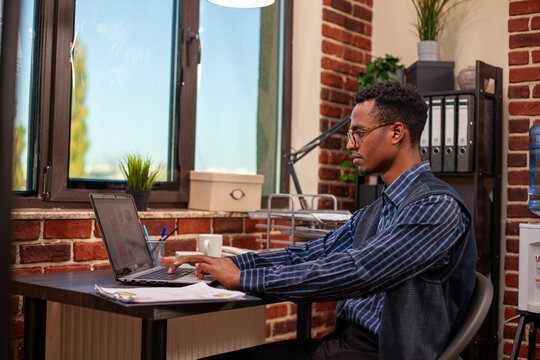 Office worker researching company data on laptop, analyzing startup information for business growth. Black man sitting at desk and using personal computer to gather insights for project development.