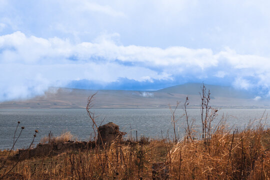 Scenic autumn landscape of Paravani Lake featuring dry golden grass, old stone ruins in the foreground, and cloudy mountains, Georgia.