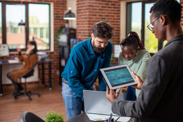 Fototapeta premium Businesswoman and colleague looking at financial data on laptop in startup office as black man holds a digital device. African american male analyst stands and reviews company annual report on tablet.