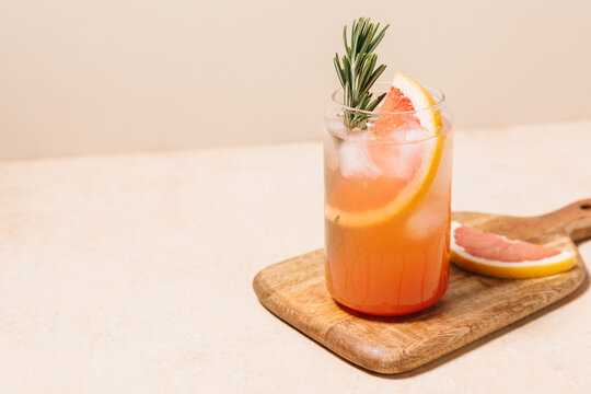 Grapefruit cocktail with rosemary garnish in a glass, served on a wooden cutting board. Vibrant, refreshing drink perfect for summer menu or cocktail recipes. Greyhound, lemonade, paloma, mocktail.