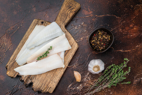 Raw cod or hake fillets with thyme on wooden board and pepper on dark textured background. Fresh pollock whitefish prepared for healthy seafood cooking.
