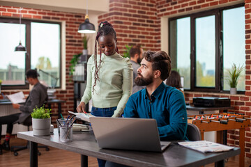 Obraz premium Black businesswoman with a clipboard discusses marketing strategy ideas with male colleague in brick wall workspace. Caucasian bearded man listens intently to female coworker at his desk in office.