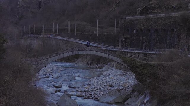 Aerial view shows a stone arch bridge over a boulder strewn river in an Italian valley, a person standing, a curving roadway, and an electrified railway at twilight.