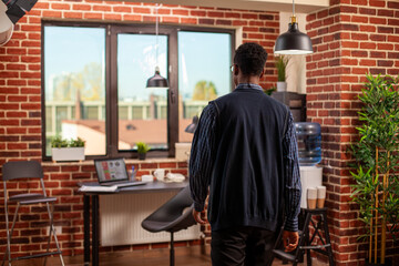 Fototapeta premium Black businessman walking into brick wall room, heading to sit at his well organized office desk. African american male manager entering modern workspace, ready for work at startup company.
