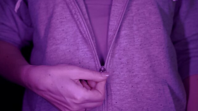 A close-up view of a female hand unzipping a grey hooded sweatshirt illuminated by dramatic purple neon lighting. Concept of casual fashion style, getting comfortable, and urban aesthetics.