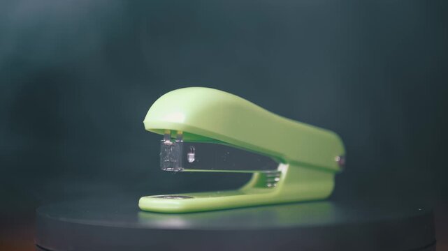 A bright green stapler stands on a dark surface while white smoke drifts through the background. Concept of office supplies, school equipment, and professional stationery organization.
