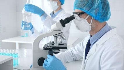 Fototapeta premium Masked scientist examining specimen under microscope while laboratory assistant handles glassware in a professional medical investigation. Health care and science concept