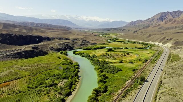 Aerial view captures peaceful Chu river winding through mountainous Boom gorge. Scenic landscape features railroad tracks, cars, and lush vegetation under clear daytime sky.