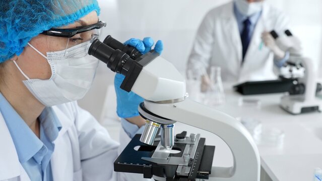 Female scientist wearing a mask and blue safety gear, using a microscope for research while a male colleague works in the background. Medicine, healthcare and science concept