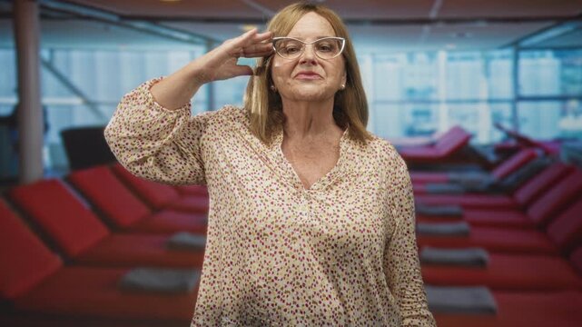 Senior hispanic blonde woman salutes with right hand to forehead and adjusts eyeglasses while standing before red lounge chairs and large windows in airport terminal; pride.