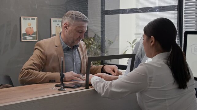 Customer reviewing documents at bank counter while employee working at computer behind glass partition