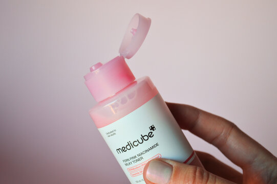 Closeup of a person's hand holding a Medicube PDRN Pink Niacinamide Milky Toner bottle with an open flip-top cap against a soft pastel background.