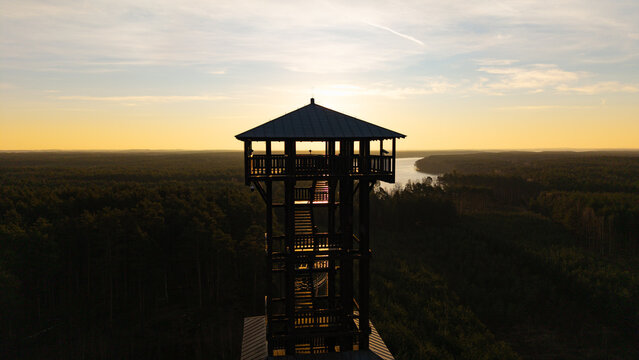 Aerial drone footage of a wooden observation tower surrounded by forest with a lake in the background at sunrise, warm morning light, peaceful nature landscape.