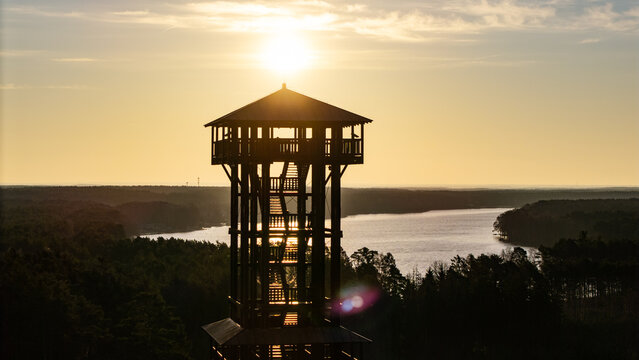 Aerial drone footage of a wooden observation tower surrounded by forest with a lake in the background at sunrise, warm morning light, peaceful nature landscape.