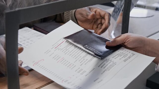 High angle close up shot of unrecognizable African American man handing documents and passport to bank teller across glass counter
