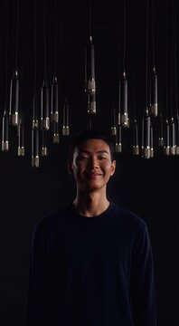 Young Asian man standing under hanging microphones, looking at the camera with a serious expression, in a dark studio setting.