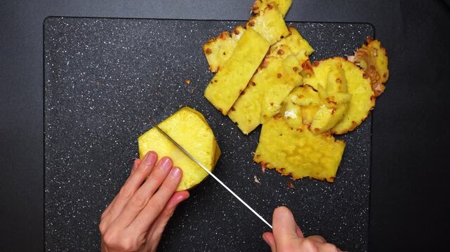 Top view of hands peeling and slicing pineapple on board. Overhead technical shot of hands using a knife to slice a fresh yellow pineapple on a black textured board.