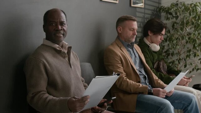 Portrait shot of African American man looking at camera while holding documents and sitting in bank waiting area with two other men