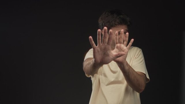 Man shielding face with both palms out and forearms exposed in studio against black backdrop; defense.