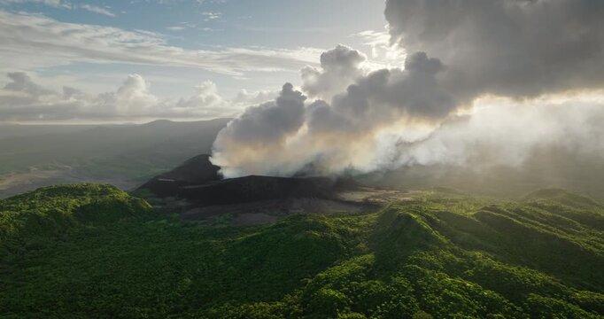 Mount Yasur volcano actively erupting in Vanuatu, releasing thick white smoke and ash into the sky, surrounded by lush green tropical vegetation on Tanna island. Aerial view drone flight panorama