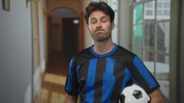 Young man holding soccer ball and shrugging in a hotel building corridor wearing blue and black striped jersey; sports resignation.