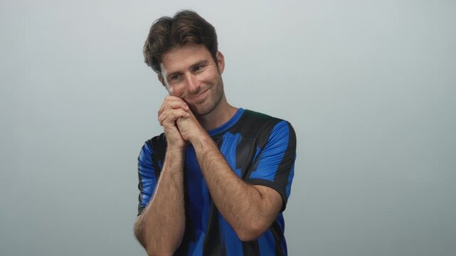 Man with hands clasped near face in a light gray studio wearing a blue and black soccer jersey; hopeful support devotion.