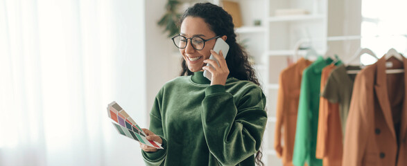 A woman stands in a clothing store talking on her phone. She holds a color palette in her hand while wearing a green sweater. © Prostock-studio