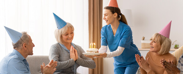 Naklejka premium Friends and caregivers gather around a woman on her birthday in a cozy room. A caregiver brings a cake with candles while others clap and smile. Moments of joy and celebration fill the space.