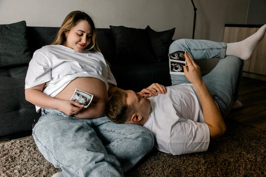 Pregnant woman and husband look at sonogram photos while relaxing on a floor carpet. The scene conveys happiness and family bonding. Suitable for parenting and maternity blogs.