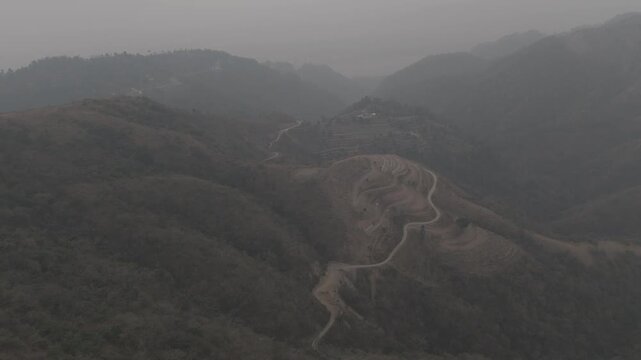 Aerial view of Dwara village near Dehradun, Uttarakhand, with a dirt path along a mountain ridge, misty hills, terrace farms, sal and pine forests, and the Song River below on a winter evening.