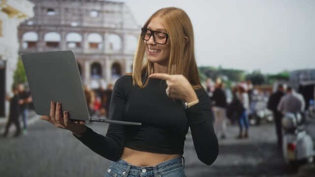Woman holds laptop and points finger to laptop on a busy street near a large building and tourist plaza; confidence success.