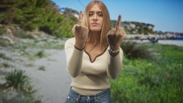 Redhead woman shows both middle fingers to camera with tense hands at a beach shoreline, wearing cream sweater and jeans, stern expression; defiance.