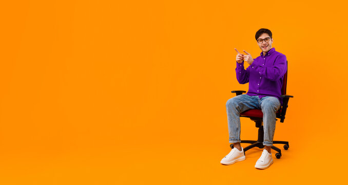 A young man sits in a black office chair in front of a bright orange background. He wears a purple shirt and jeans and points with a smile. The scene has a fun and casual vibe.