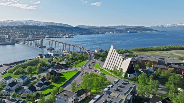 Drone aerial flying toward the Arctic Cathedral and Troms&oslash; Bridge in Troms&oslash;, Norway. Iconic white church stands near the shoreline with Arctic fjord and mountains in the background.