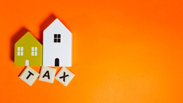 Two miniature model houses are placed above letter tiles spelling "TAX" on a vibrant orange surface, symbolizing property taxes, real estate liabilities, and the financial costs of homeownership.