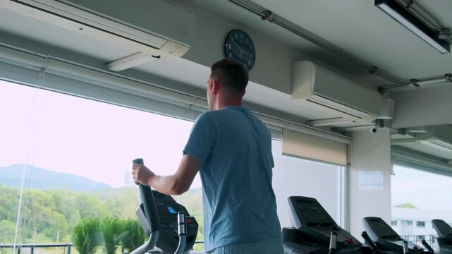 Man training on elliptical trainer in modern gym near large windows with a view of greenery. Fitness and routine, focusing on cardio exercise and active lifestyle in indoor training environment