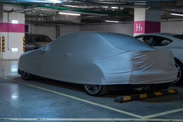 Obraz premium Mysterious luxury car, concealed beneath light grey protective cover, securely parked in dimly lit, modern underground garage with concrete pillars and visible utilities