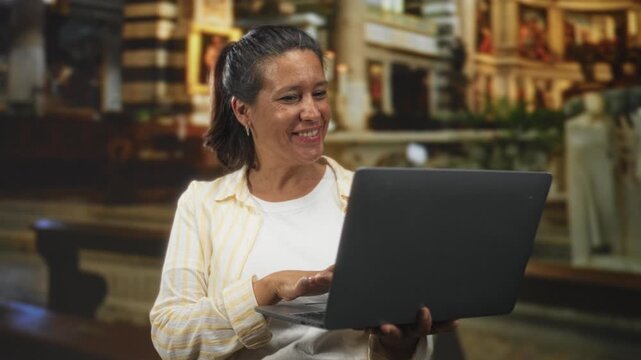Woman holding laptop and tapping the keyboard with her right hand in an ornate church interior, smiling while looking at the screen; joy.
