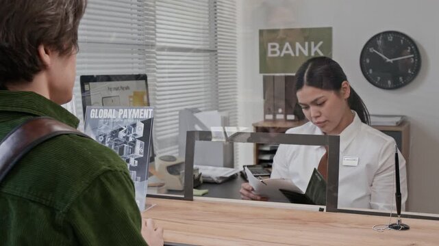 Over the shoulder shot of bank teller handling paperwork while assisting young man at modern bank