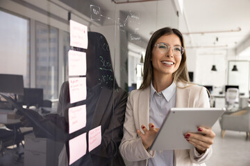 Happy businesswoman, project coordinator or marketing staff member holds tablet device stands near of wall with attached sticky notes smiling, reviewing sales results, enjoy workflow, chatting online © fizkes