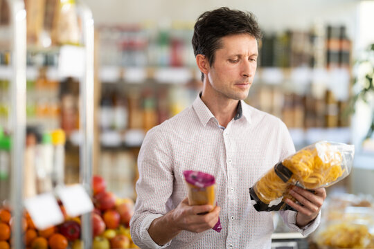 Man in light shirt took some varieties of pasta from shelf. Client compare pasta noodles items package. Customer examines contents of package, choose noodles garnish product