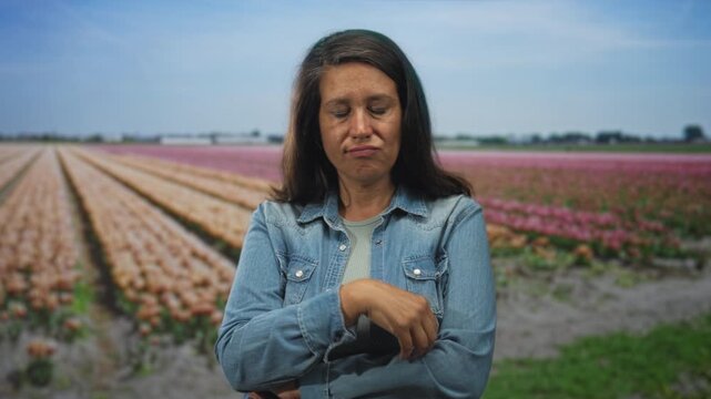 Woman, hispanic middle age brunette, wearing denim jacket with folded arms and raised hand near chin in a studio tulip field backdrop; quiet skepticism.