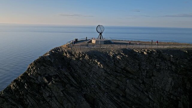 Drone aerial revealing the North Cape monument on the edge of towering Arctic cliffs in Norway. Dramatic rocky coastline meets the open Barents Sea.