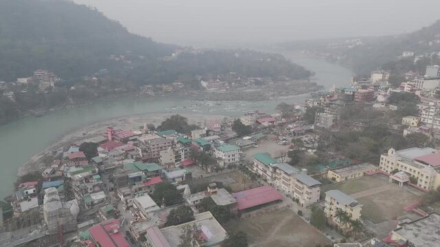 Aerial view of Lakshman Jhula in Rishikesh, Uttarakhand, with the Ganga River flowing below, and temples, houses, and buildings along the riverbank against misty mountains on a winter morning.