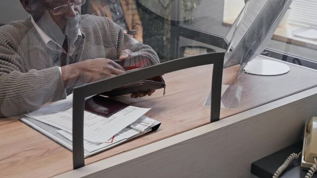 High angle shot of senior African American man handing cash money to bank teller through glass counter
